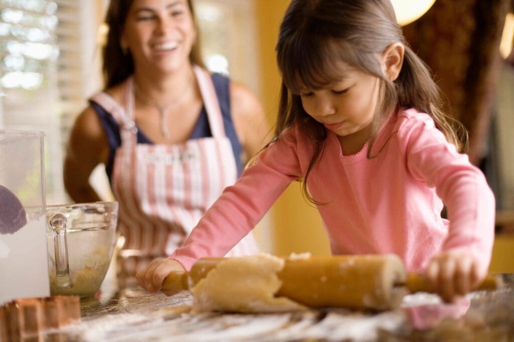 Child rolling dough, smiling woman in background.