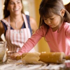 Child rolling dough, smiling woman in background.